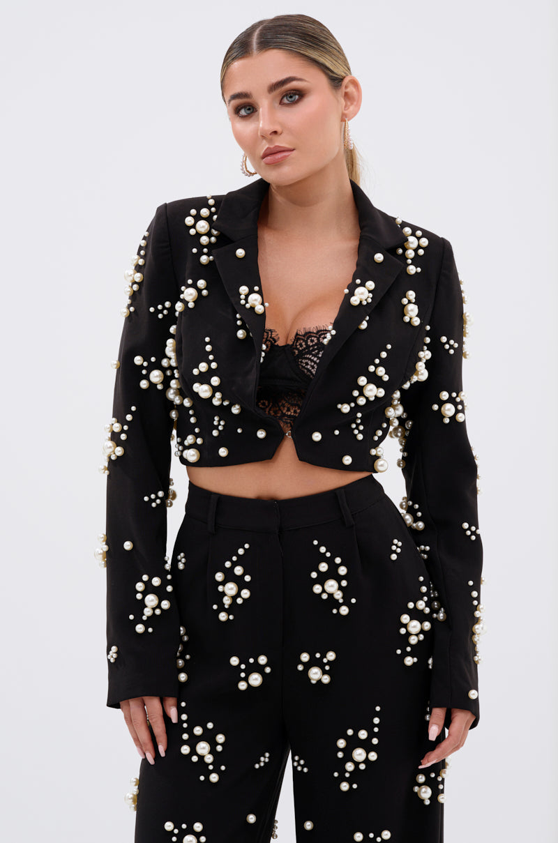 A woman stands against a plain background wearing the OLD FLAME EMBELLISHED BLAZER IN BLACK with faux pearl details, paired with matching high-waisted pants and a black lace bralette visible beneath her blazer. Her hair is pulled back for a chic look.