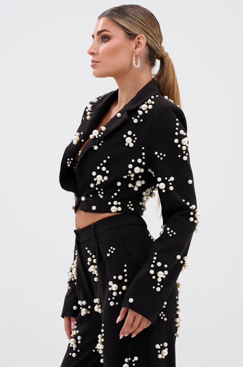 Woman in profile wears the OLD FLAME EMBELLISHED BLAZER IN BLACK with pearl clusters, paired with matching high-waisted pants. Her long straight hair is styled in a low ponytail, and she finishes the look with hoop earrings.