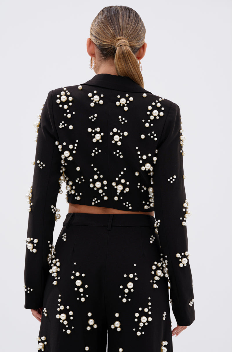 A woman with a low ponytail is seen from behind in the OLD FLAME EMBELLISHED BLAZER IN BLACK and matching pants, both adorned with clusters of white pearls, against a plain white background.