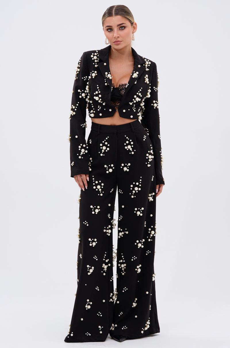 A woman wears the OLD FLAME EMBELLISHED BLAZER IN BLACK with wide-leg pants and a black lace bralette, featuring pearl-like embellishments, set against a plain white background for a chic look.