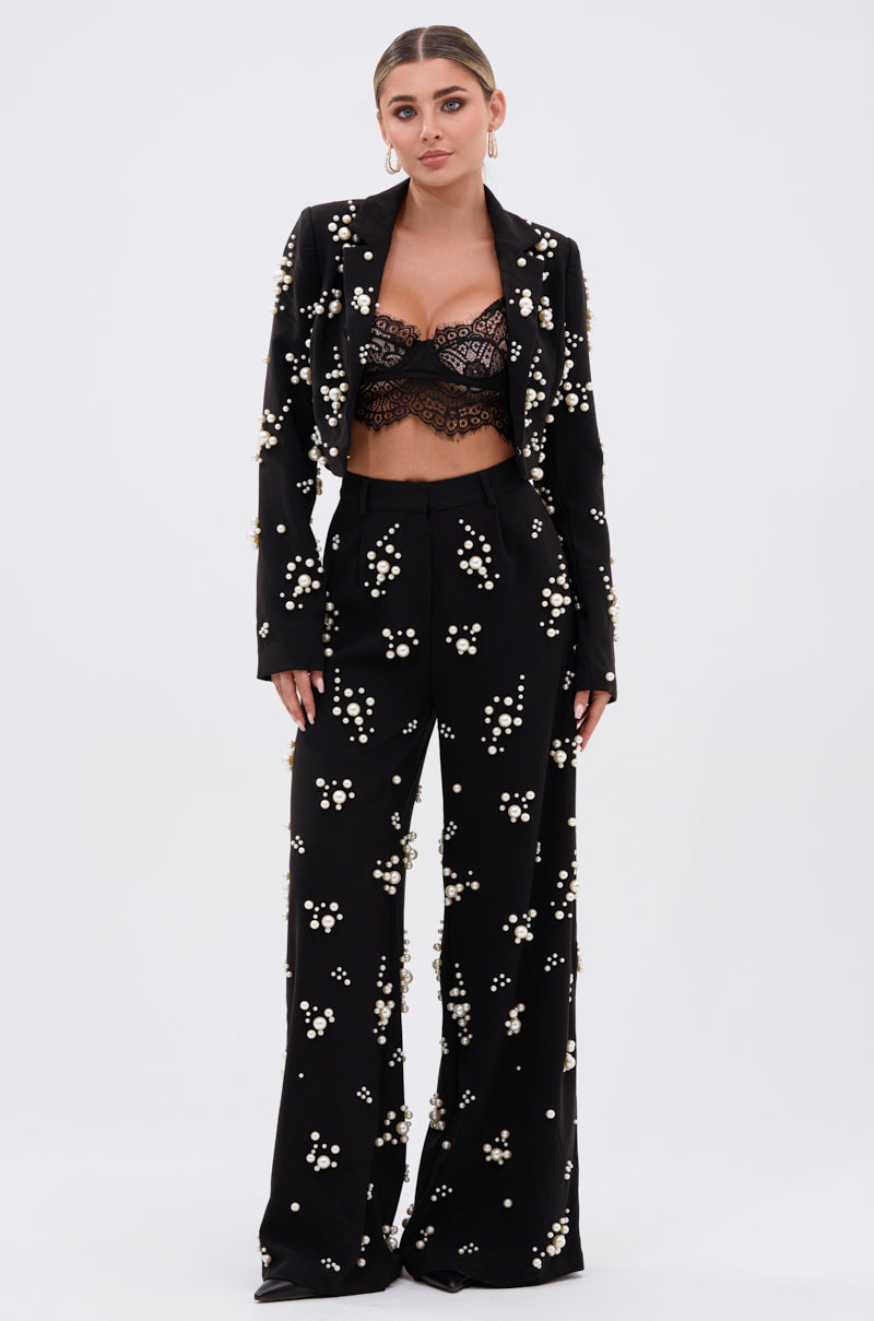 A woman with her hair pulled back and a neutral expression faces forward, wearing the OLD FLAME EMBELLISHED BLAZER IN BLACK over a black lace bralette, set against a plain light background.