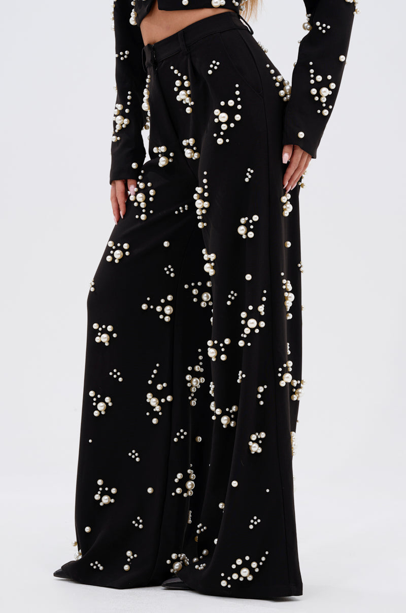 A person in the OLD FLAME EMBELLISHED TROUSER IN BLACK—high-rise, wide-leg pants with faux pearl details—paired with a matching long-sleeve top, stands against a plain light background.