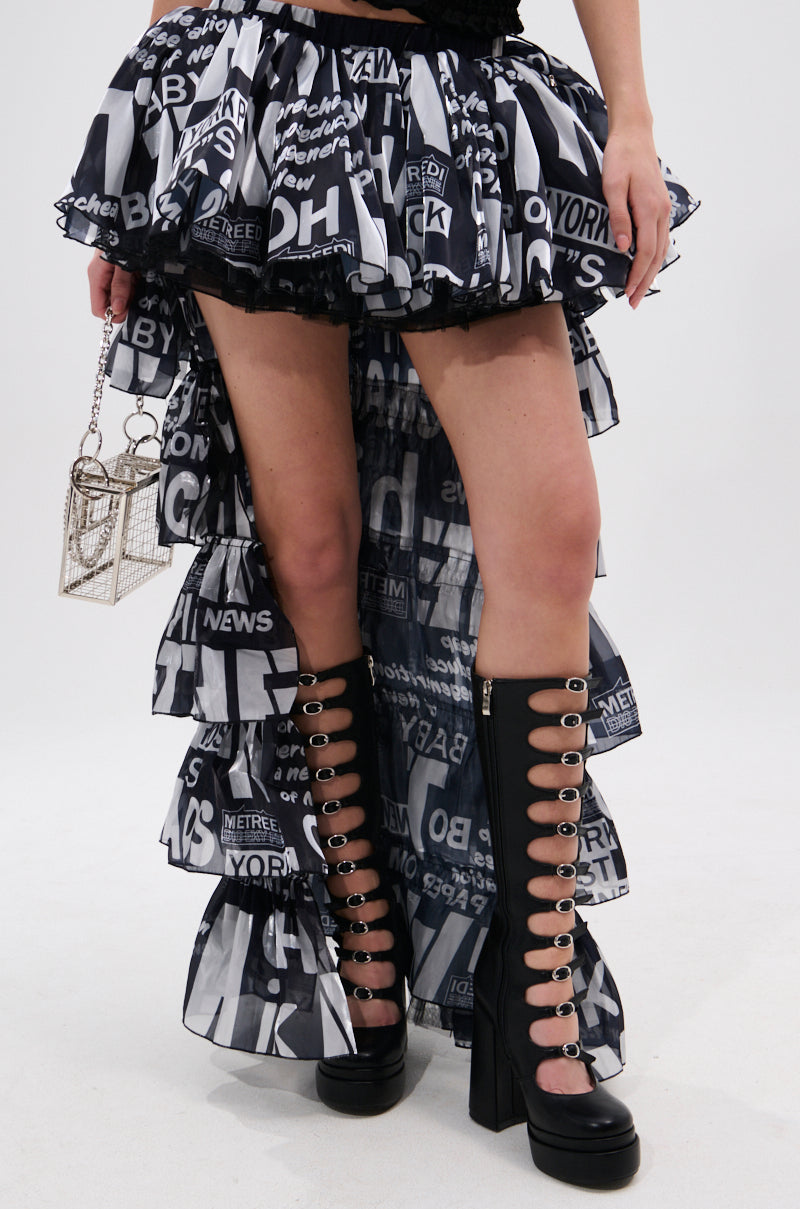 A person models the CRAZY FOR YOU HIGH LOW GRAPHIC SKIRT, a ruffled black-and-white skirt with a newspaper-style print and elastic waistband, styled with knee-high black platform boots and a boxy handbag against a plain white background.