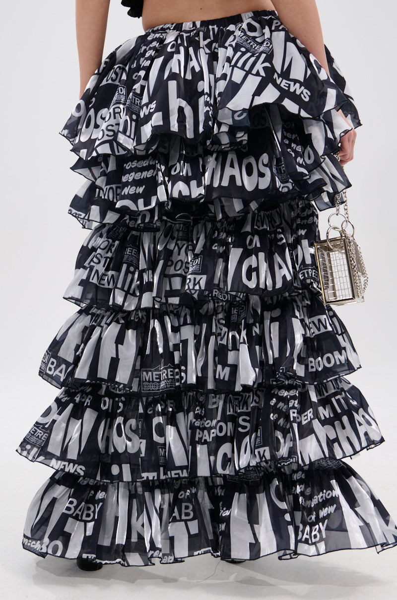 A person wearing the CRAZY FOR YOU HIGH LOW GRAPHIC SKIRT, featuring black-and-white tiered ruffles, an elasticized waistband, and bold newspaper-style prints, is shown from the waist down holding a small silver handbag.