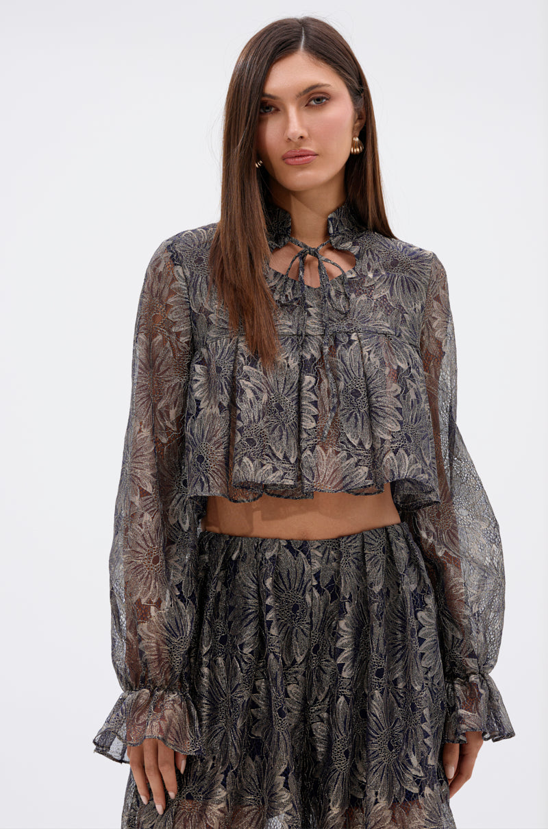 A woman with straight brown hair wears the BEST GUEST LACE CROP BLOUSE, a sheer navy long-sleeved floral top with a tied neckline, paired with a matching high-waisted skirt, standing against a plain light background.