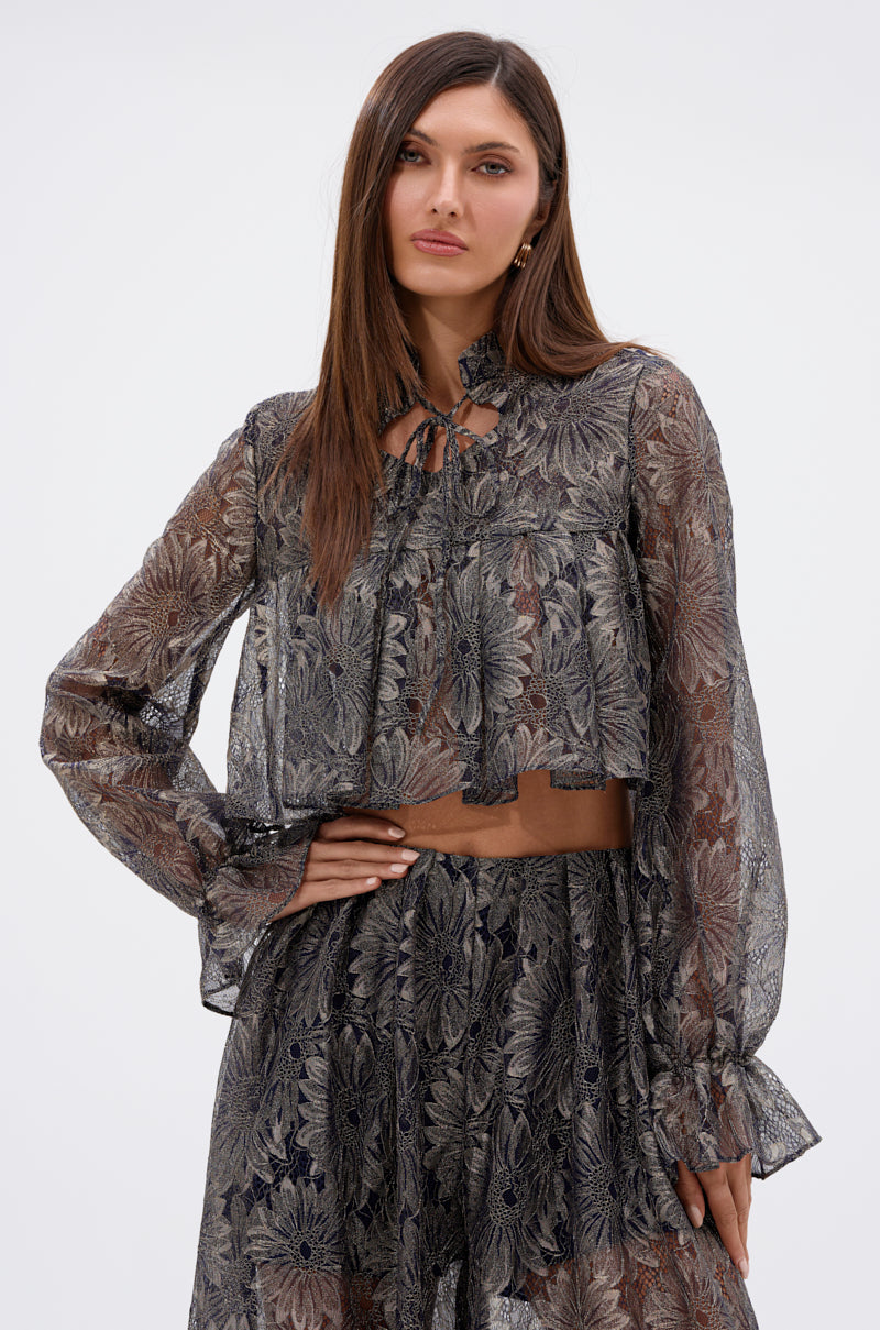A woman with long brown hair wears the BEST GUEST LACE CROP BLOUSE, a sheer, long-sleeve lace crop top paired with a matching high-waisted skirt featuring a metallic floral overlay, posing against a plain light background. 1