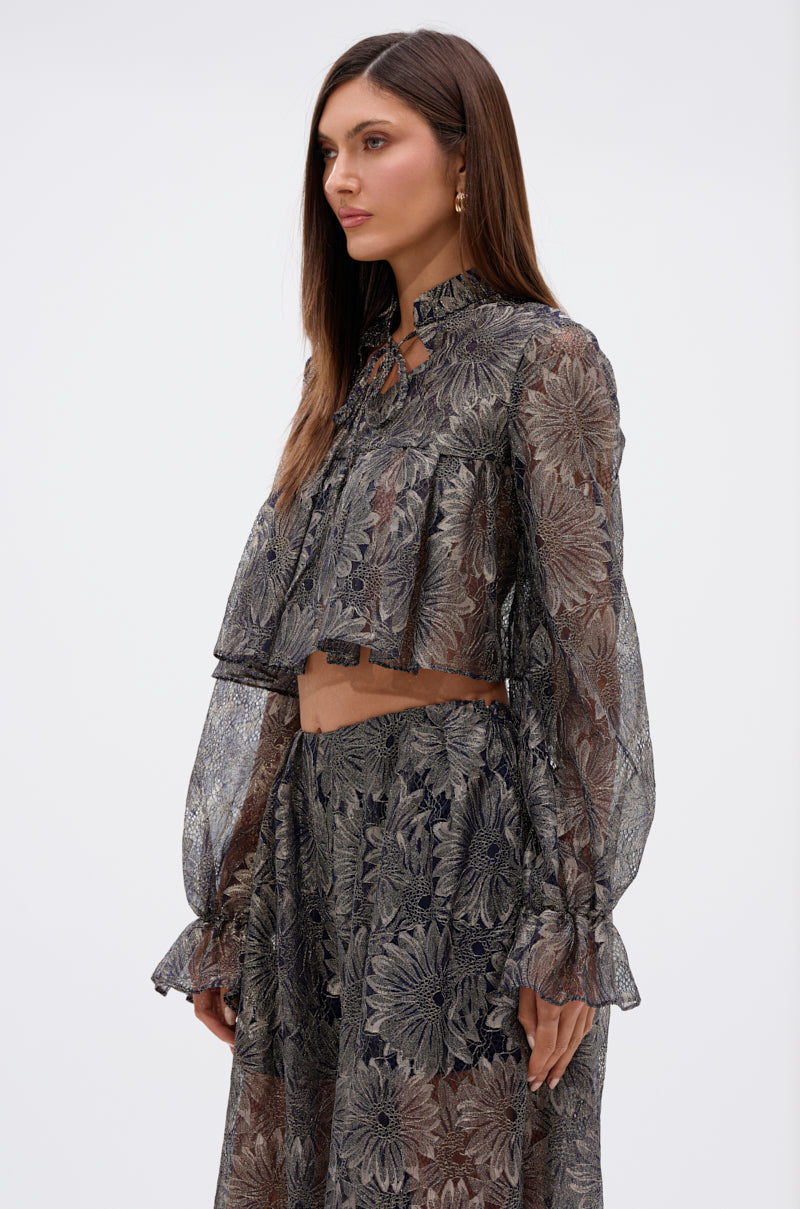 A woman stands in profile wearing the BEST GUEST LACE CROP BLOUSE—a sheer navy, long-sleeve, floral lace top with a ruffled high neck—paired with a matching skirt. Her brown hair is straight and loose as she gazes forward neutrally. 2