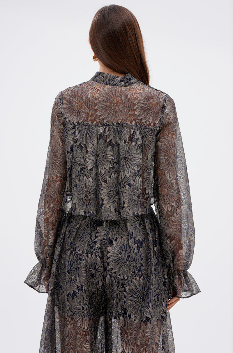 A woman with long brown hair faces away, wearing the BEST GUEST LACE CROP BLOUSE—a sheer, long-sleeved navy top with an intricate gray and navy floral pattern—paired with a matching skirt against a plain background. 3