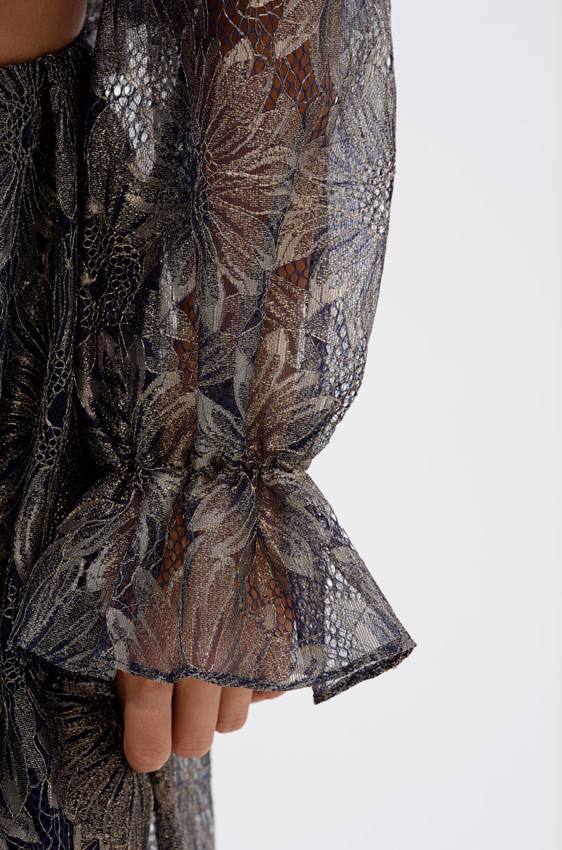 A close-up of a hand and arm wearing the BEST GUEST LACE CROP BLOUSE, a sheer long-sleeved top with ruffled cuffs and metallic floral blue and gold details, reminiscent of navy lace. 6