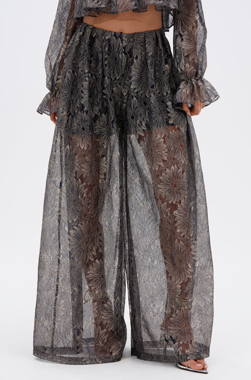 Wearing the BEST GUEST LACE PALAZZO PANT, featuring high-rise wide legs and a sheer black metallic floral overlay with visible shorts, paired with a matching sheer long-sleeved top. The person stands against a plain white background.