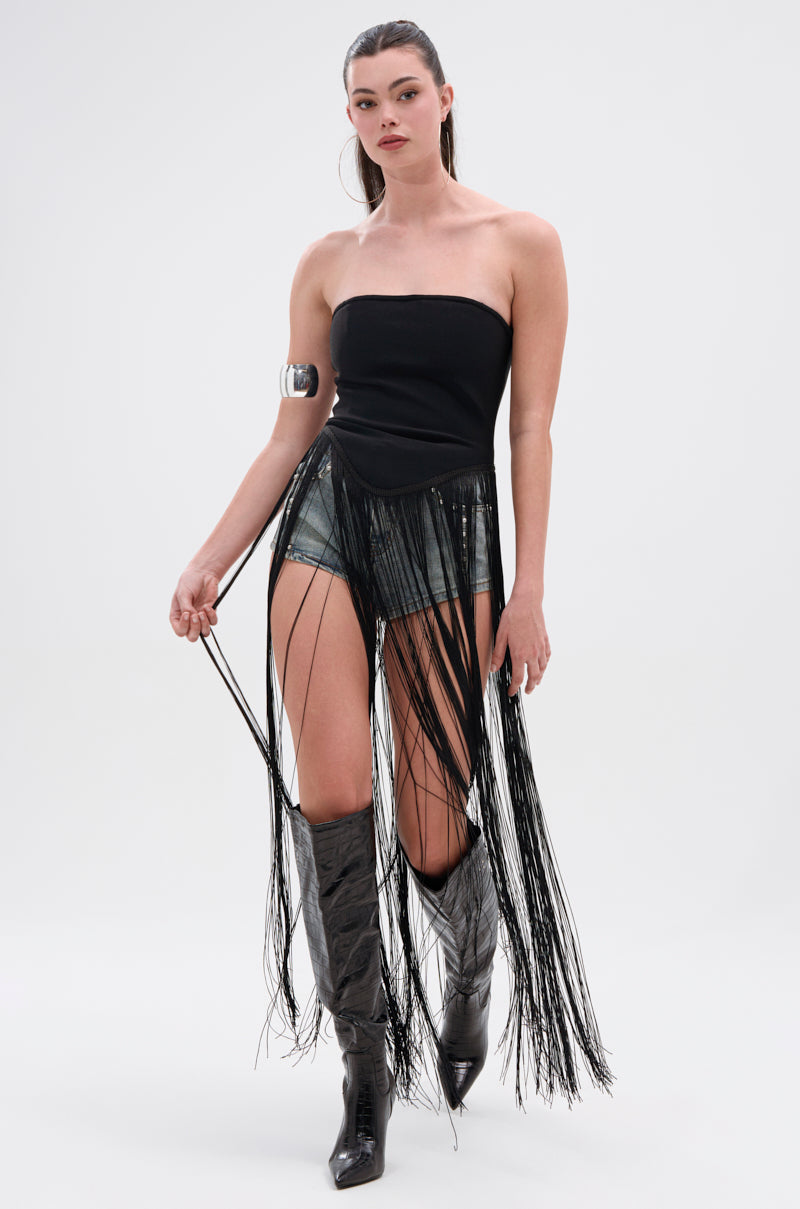A woman stands against a plain background wearing the GONNA LOVE ME FRINGE TOP, a black strapless style with long fringe, paired with denim shorts, shiny knee-high boots with a metallic zipper, and a metallic arm cuff. Her hair is tied back.