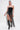 A woman stands against a plain background wearing the GONNA LOVE ME FRINGE TOP, a black strapless style with long fringe, paired with denim shorts, shiny knee-high boots with a metallic zipper, and a metallic arm cuff. Her hair is tied back.