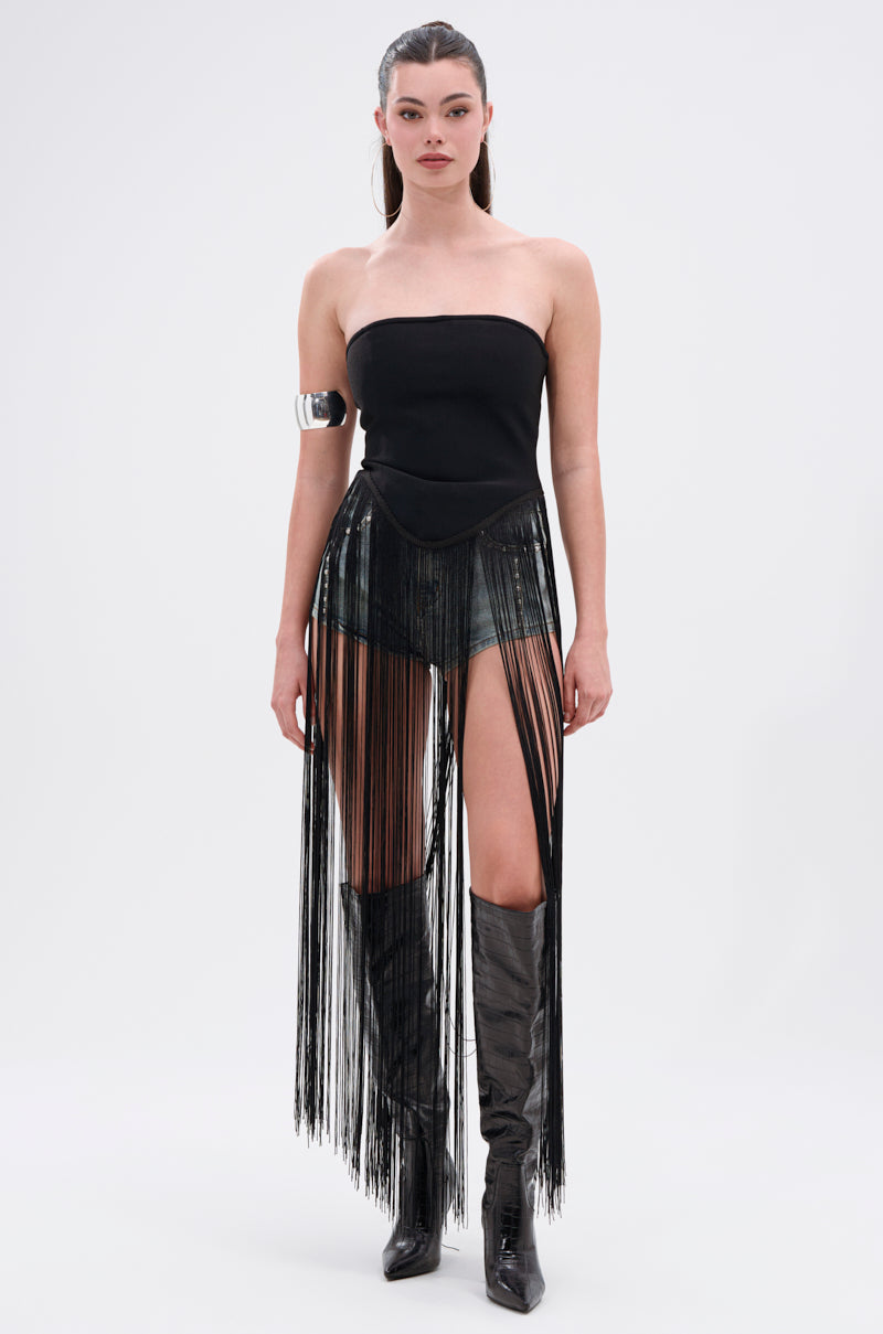 A woman stands facing forward, wearing the GONNA LOVE ME FRINGE TOP, short denim shorts, long black fringe skirt, metallic arm cuff, shiny black knee-high boots. Her hair is slicked back and she has dark lipstick.