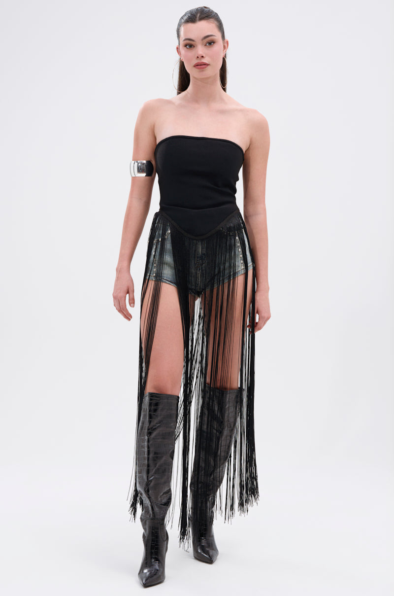 A woman stands against a plain white background, wearing the GONNA LOVE ME FRINGE TOP—a strapless black fringe bodysuit with high-waisted shorts—paired with metallic knee-high boots, a silver armband, and her hair in a sleek high ponytail.