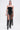A woman stands against a plain white background, wearing the GONNA LOVE ME FRINGE TOP—a strapless black fringe bodysuit with high-waisted shorts—paired with metallic knee-high boots, a silver armband, and her hair in a sleek high ponytail.