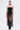A woman faces forward in the GONNA LOVE ME FRINGE TOP, styled with metallic shorts, a long black fringe skirt, knee-high boots, hoop earrings, a metal armband, and slicked-back hair against a plain white background.