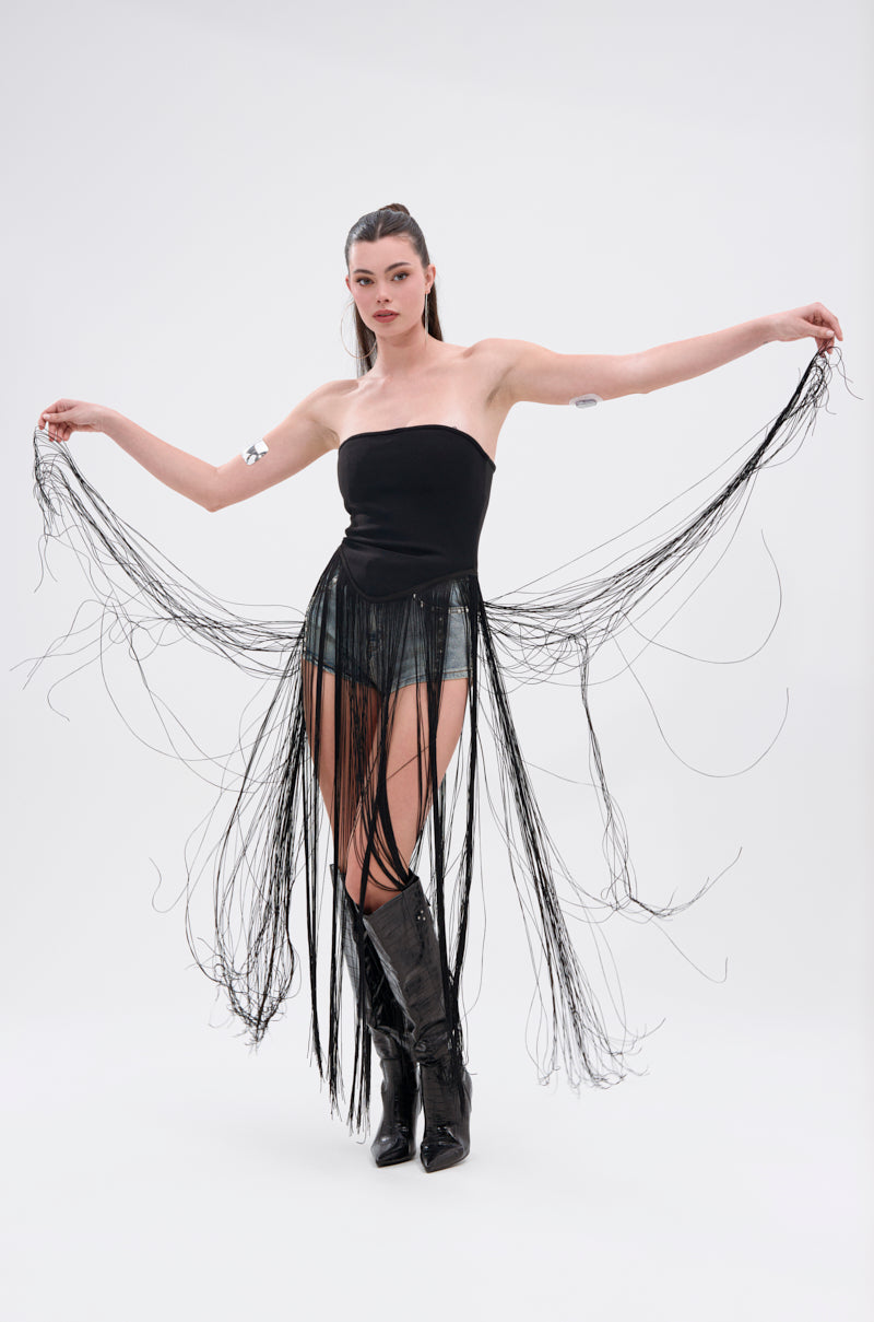 A woman stands with arms outstretched, wearing the GONNA LOVE ME FRINGE TOP, black shorts, tall black boots, and a flowing fringed black skirt. The background is plain white.