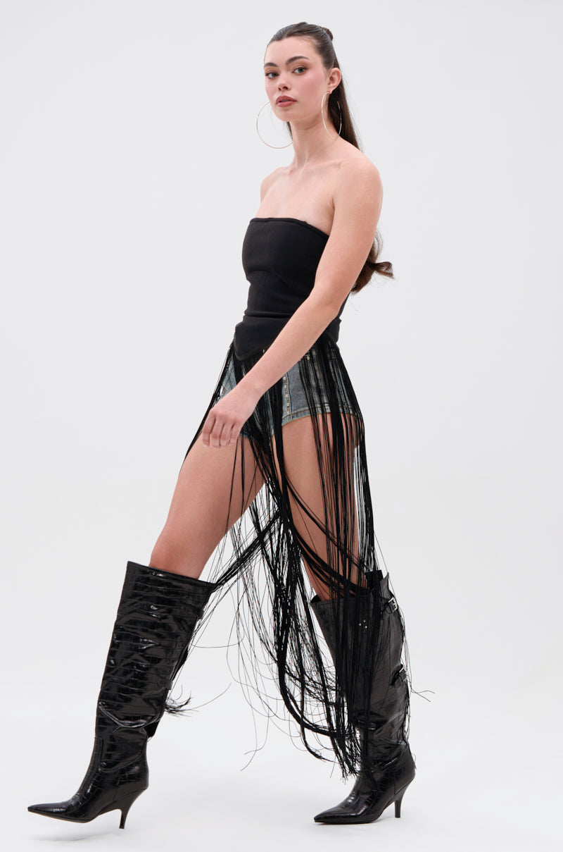 A woman models the GONNA LOVE ME FRINGE TOP—black, strapless with metallic zipper—paired with denim shorts, long black fringe skirt overlay, tall shiny boots, and hoop earrings. Her hair is tied back against a plain white background.
