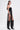 A woman stands in profile wearing the GONNA LOVE ME FRINGE TOP, metallic shorts, a long black fringe skirt, knee-high black boots, and a chunky metallic armband. Her long hair is pulled back as she poses against a plain white background.