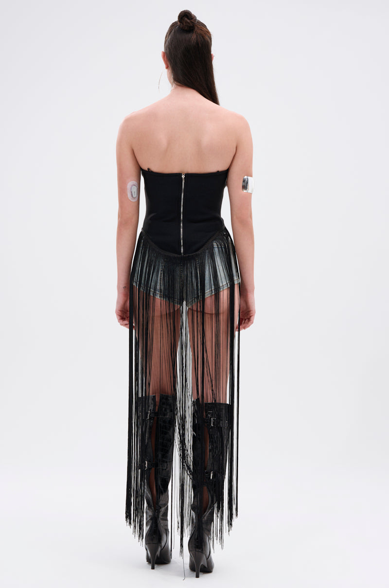 A person stands facing away, wearing the GONNA LOVE ME FRINGE TOP along with metallic shorts, a long black fringe skirt, tall black boots, and a strapless black top with zipper. Their long hair is tied up and medical patches cover their upper arms.