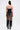 A person stands facing away, wearing the GONNA LOVE ME FRINGE TOP along with metallic shorts, a long black fringe skirt, tall black boots, and a strapless black top with zipper. Their long hair is tied up and medical patches cover their upper arms.