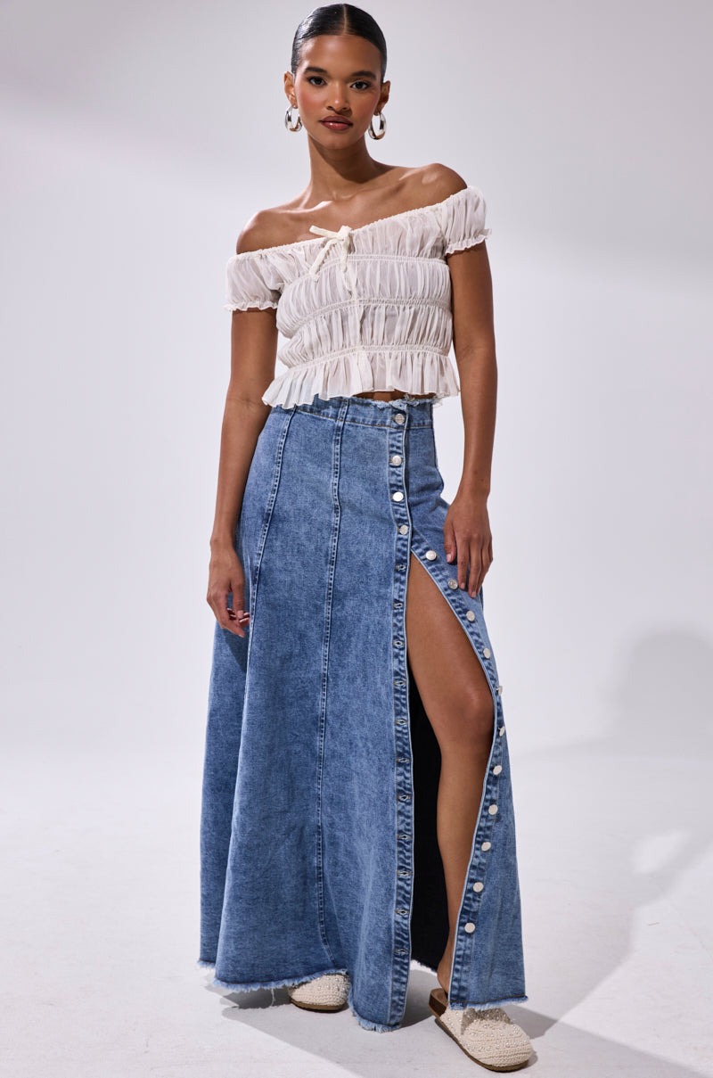 A woman poses in a white off-the-shoulder ruffled crop top and the TIFFANY BLUE DENIM MAXI SKIRT, featuring a front slit, button details, and distressed hem. She accessorizes with hoop earrings and slip-on shoes against a plain light background.