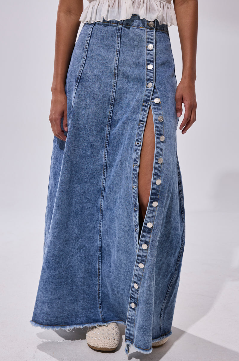A woman wears the TIFFANY BLUE DENIM MAXI SKIRT, featuring a high-rise fit, front slit, and silver buttons, styled with a white top and cream textured shoes.