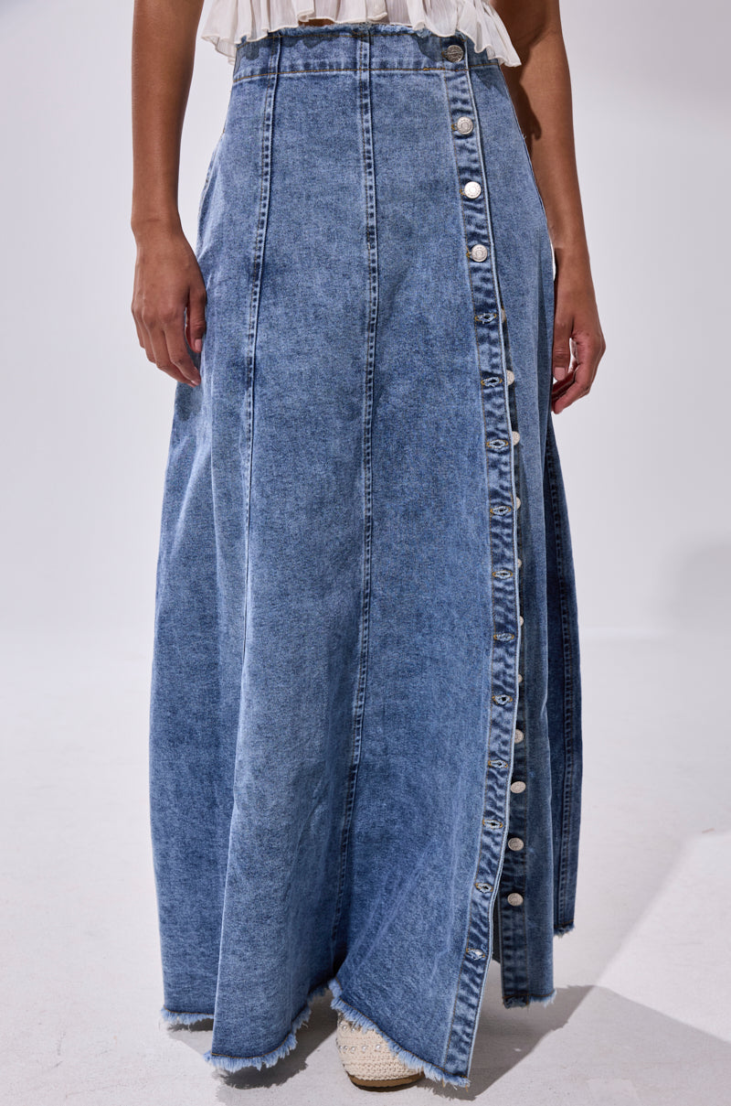 A person wears the TIFFANY BLUE DENIM MAXI SKIRT with a high-rise fit, frayed hem, and diagonal buttons, styled with a light-colored top and woven shoes.