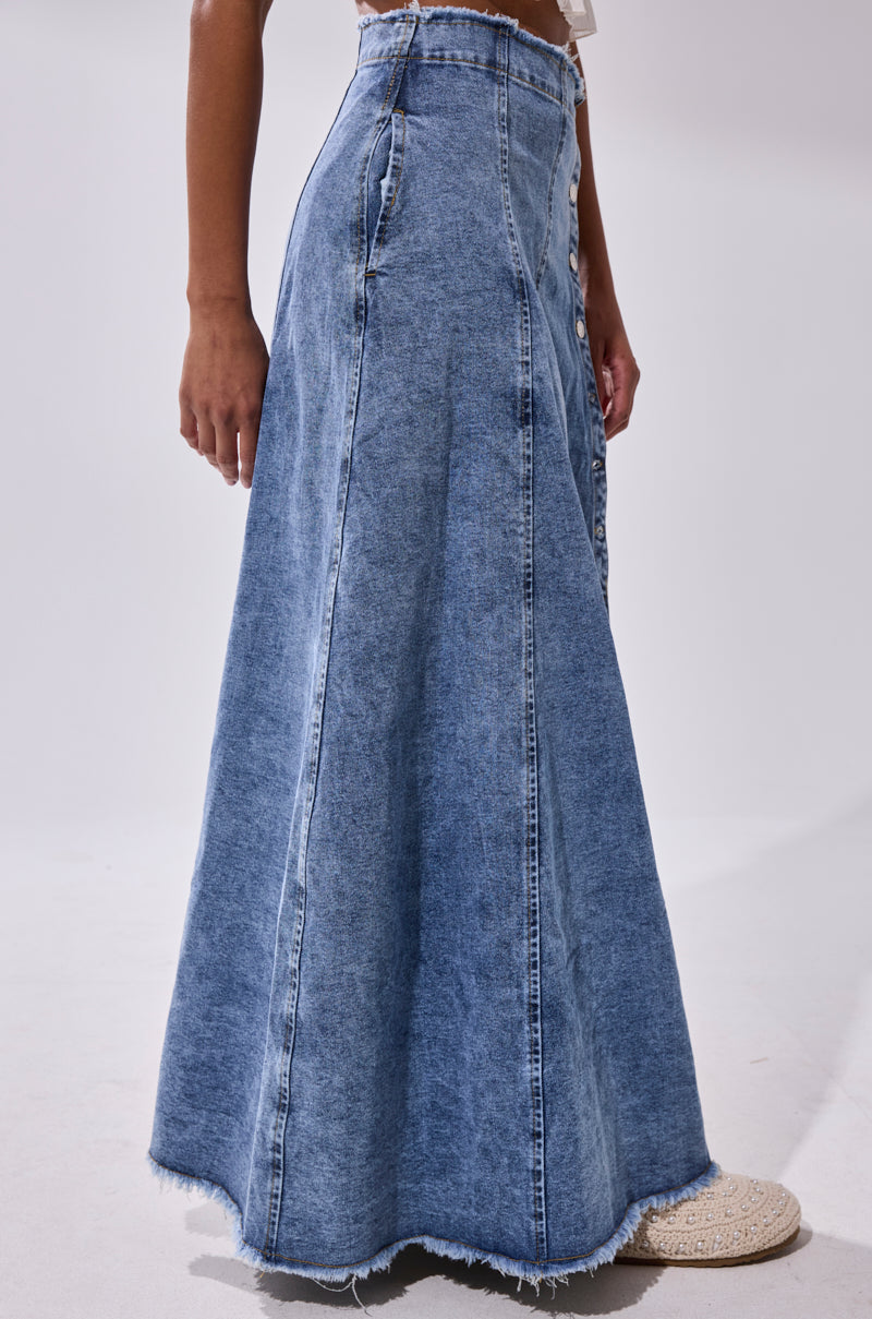 A person wears the TIFFANY BLUE DENIM MAXI SKIRT, a high-rise A-line style with vertical seams and a frayed hem, paired with cream shoes. The side view is against a plain background.