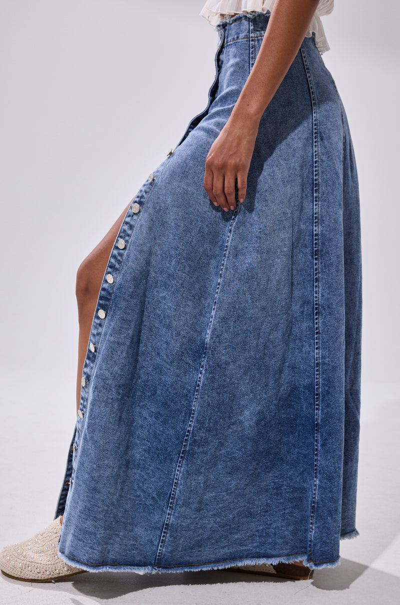 A person wearing the TIFFANY BLUE DENIM MAXI SKIRT, a high-rise button-front denim maxi skirt with a front slit, paired with white woven shoes.