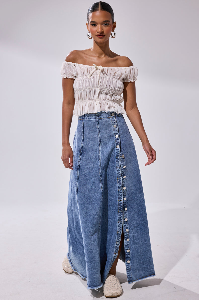Woman wears an off-shoulder white ruched top and the TIFFANY BLUE DENIM MAXI SKIRT, featuring a high-rise fit, button-front detail, and front slit, standing against a plain white background.