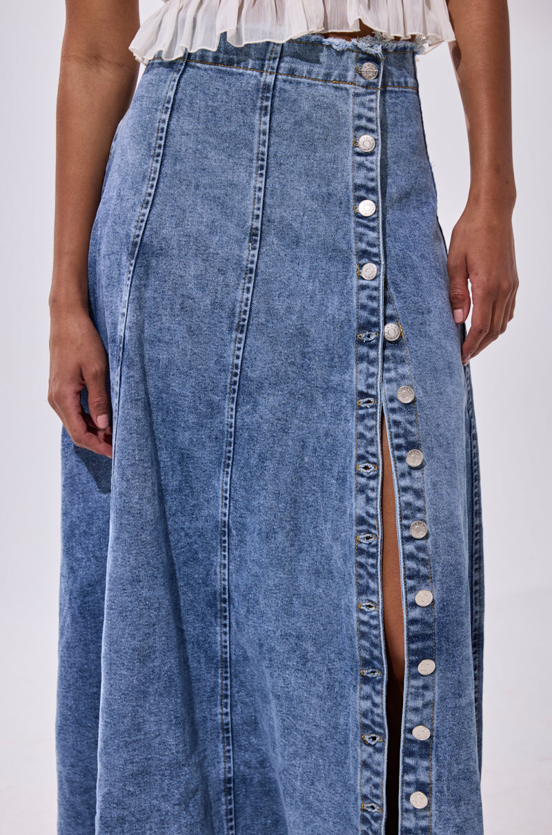 A person is seen from the torso down, wearing the TIFFANY BLUE DENIM MAXI SKIRT with vertical seams, a front silver button slit, a subtle distressed hem, and paired with a white ruffled top.