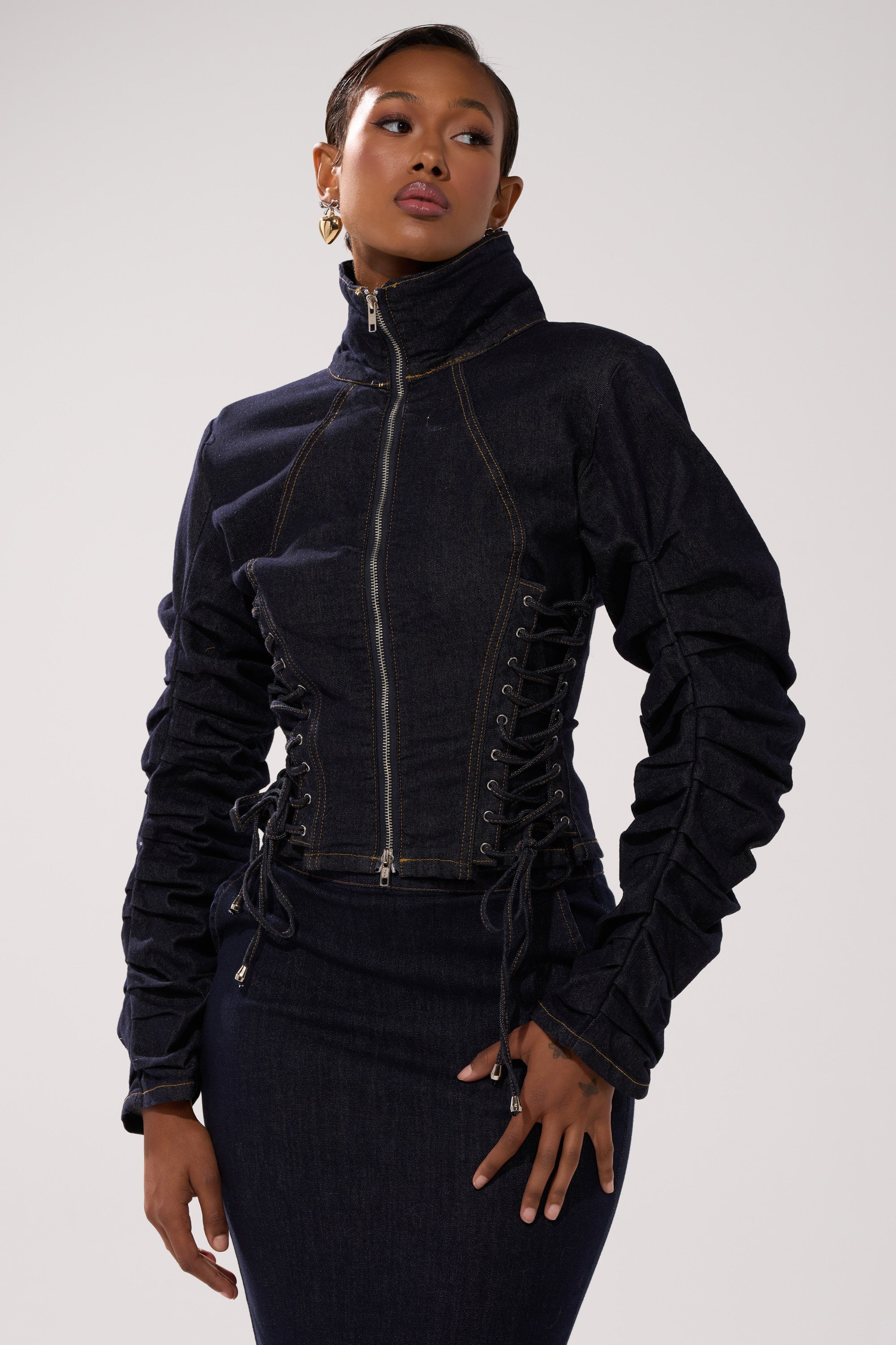 A woman poses confidently against a plain background, wearing the STRICTLY BUSINESS DENIM BLOUSE—a dark-wash jacket with a high collar, front zipper, lace-up sides, and ruched sleeves—paired with a matching skirt.