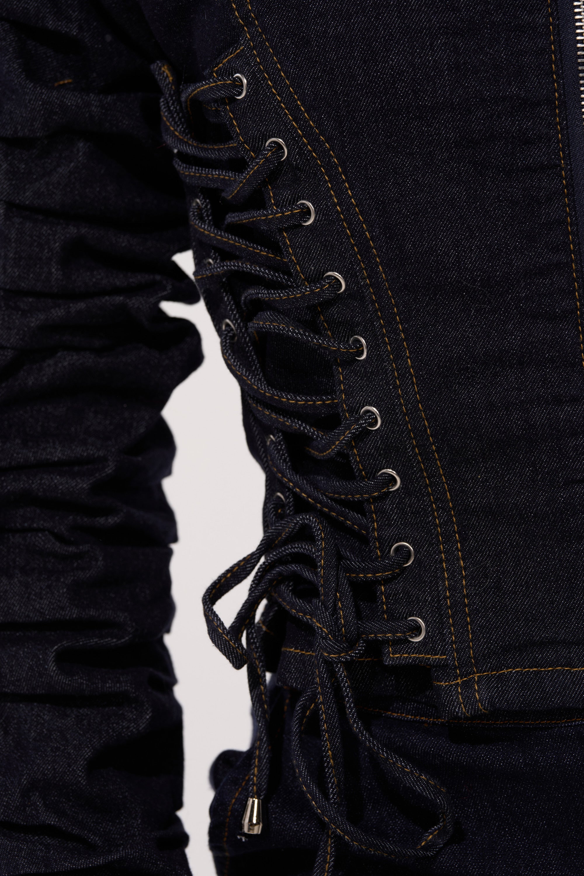 The STRICTLY BUSINESS DENIM BLOUSE is a dark-wash, cropped denim jacket detailed with yellow stitching, silver eyelets, and crisscross laces tied in a bow along the side. Its ruched sleeves add unique texture to this stylish top.