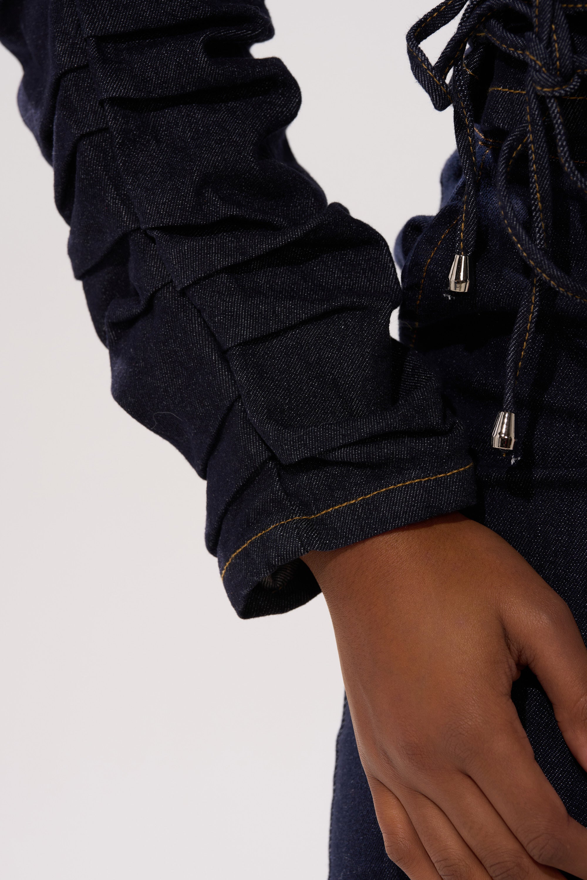 A close-up of a hand at their side, wearing the STRICTLY BUSINESS DENIM BLOUSE in dark-wash denim with ruched details and yellow stitching on the sleeve, set against a plain, light background.