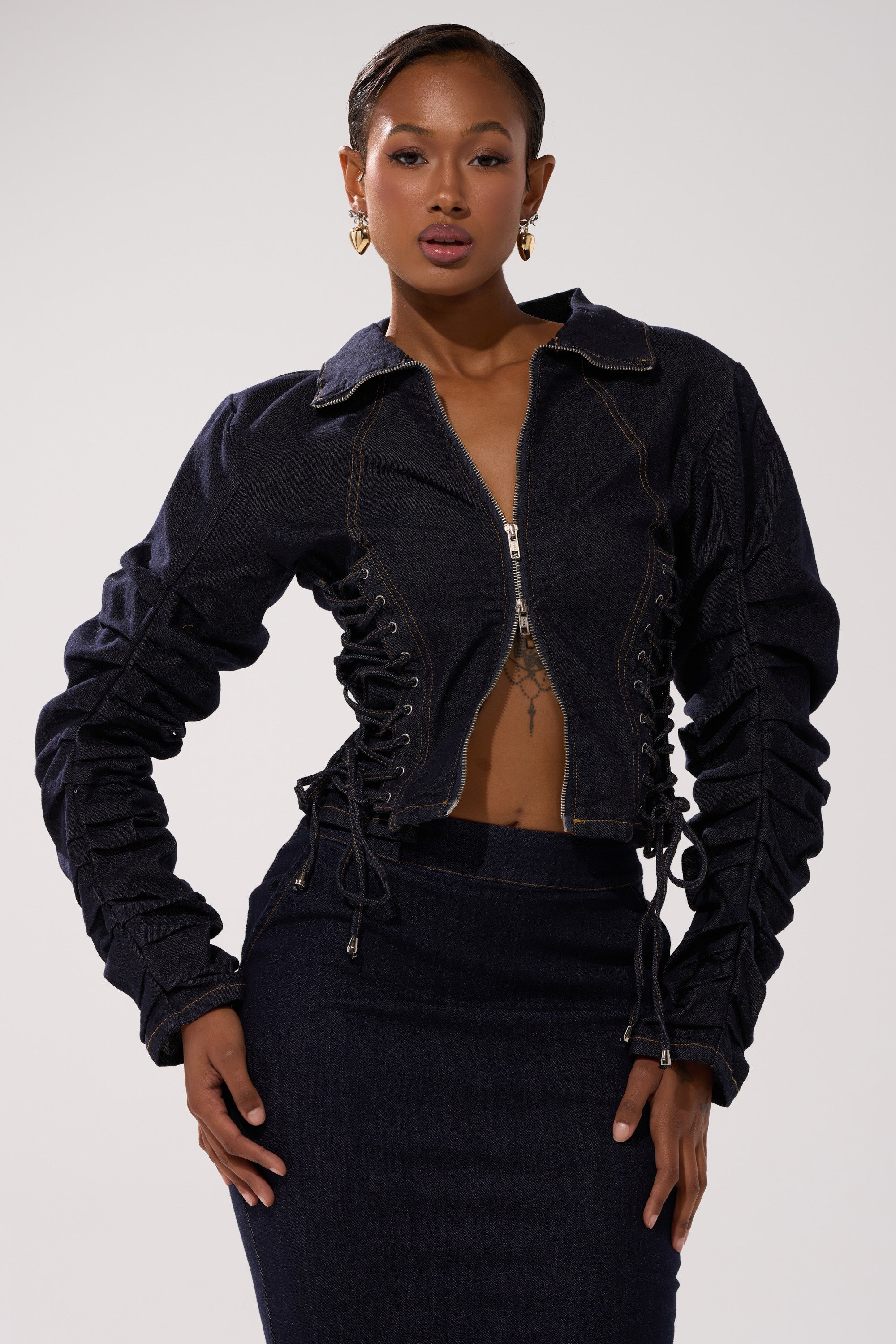A woman poses confidently in the STRICTLY BUSINESS DENIM BLOUSE, a structured dark-wash denim jacket with front zipper, ruched sleeves, and lace-up details, paired with a matching skirt against a light background.