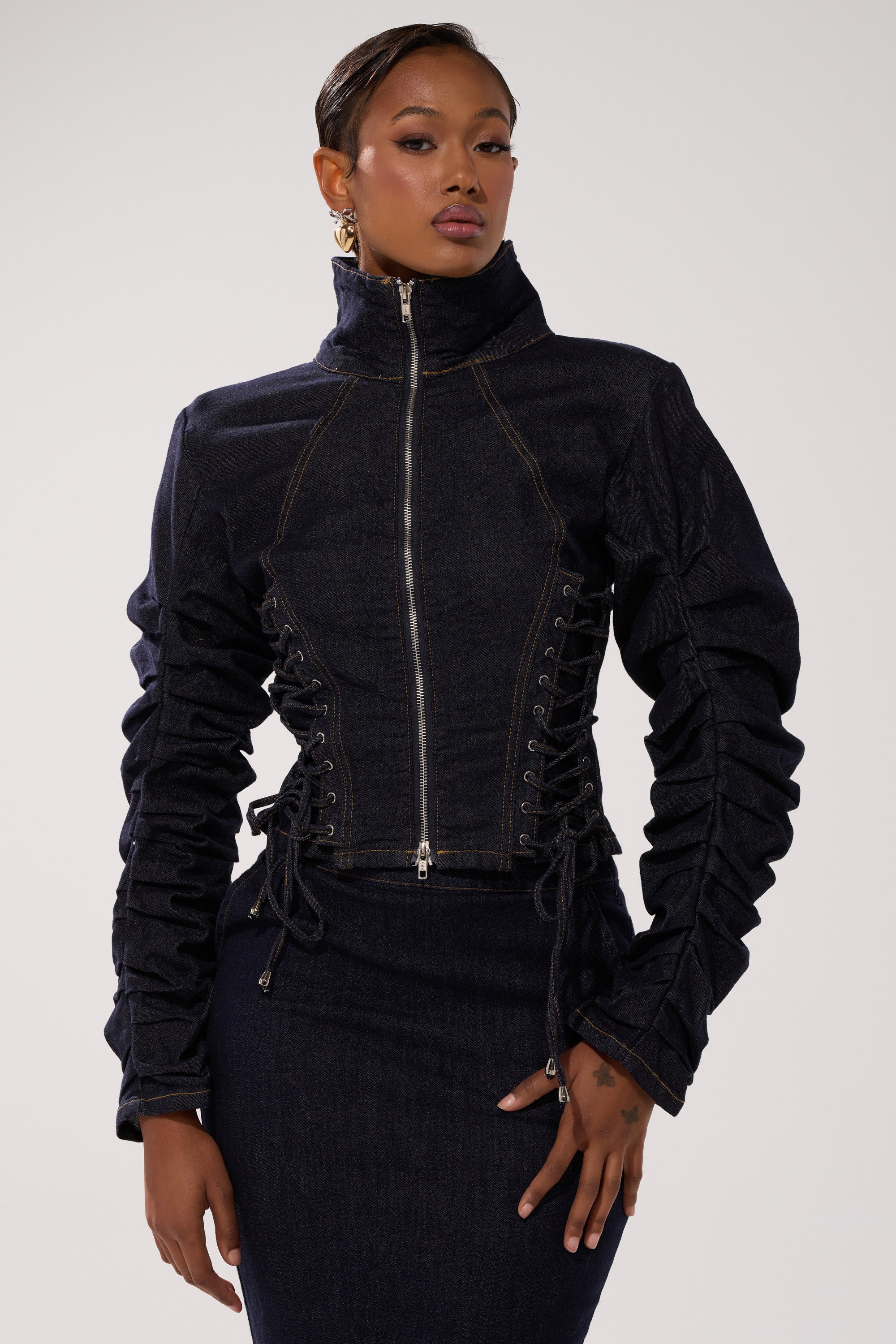 A woman poses against a plain light background wearing the STRICTLY BUSINESS DENIM BLOUSE, a dark denim zip-up cropped top with corset-style side lacing and ruched sleeves, paired with a matching dark denim skirt.