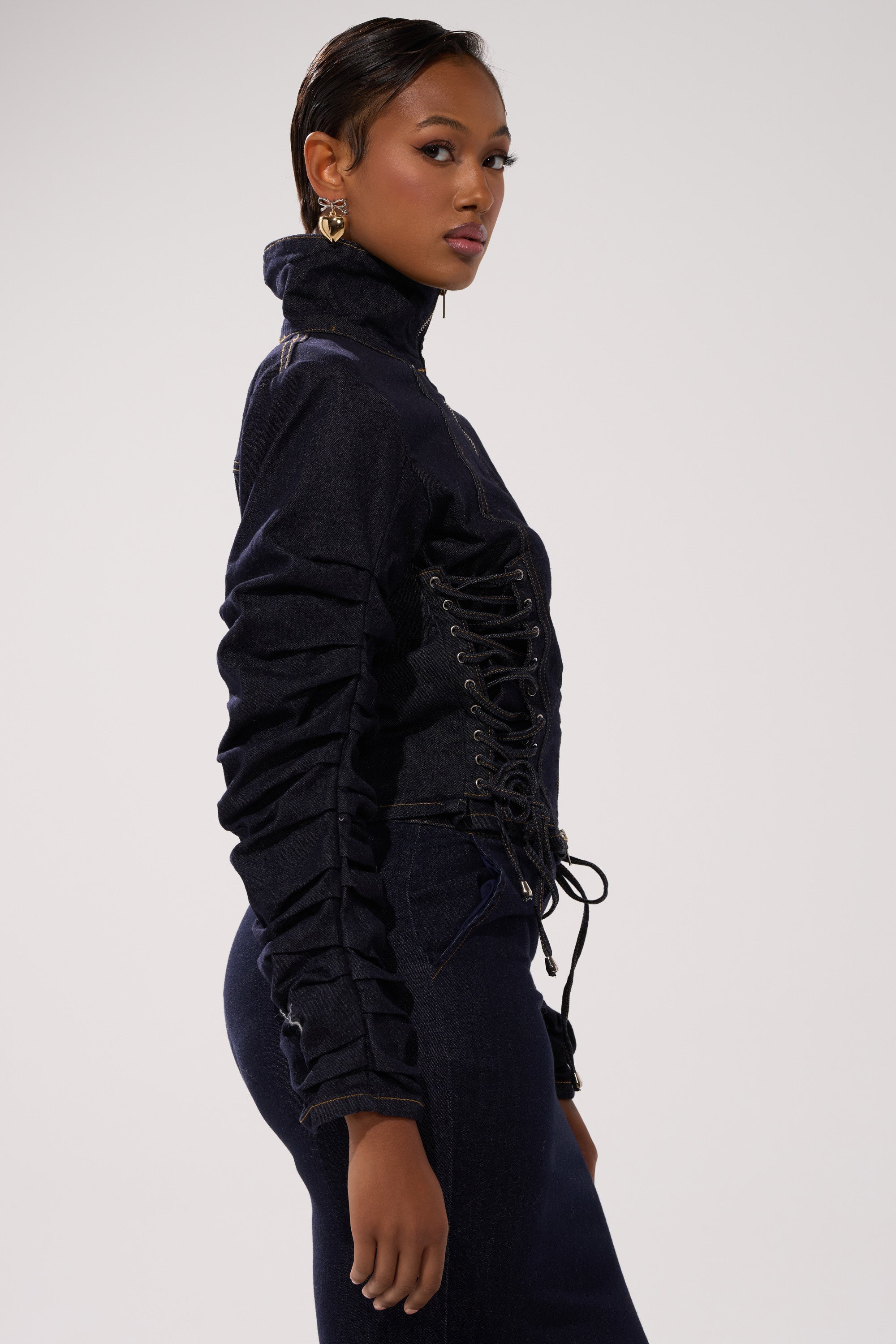 A woman stands in profile against a light background, wearing the STRICTLY BUSINESS DENIM BLOUSE with lace-up side detailing and matching pants. She has slicked-back hair and large, round earrings for accessories.