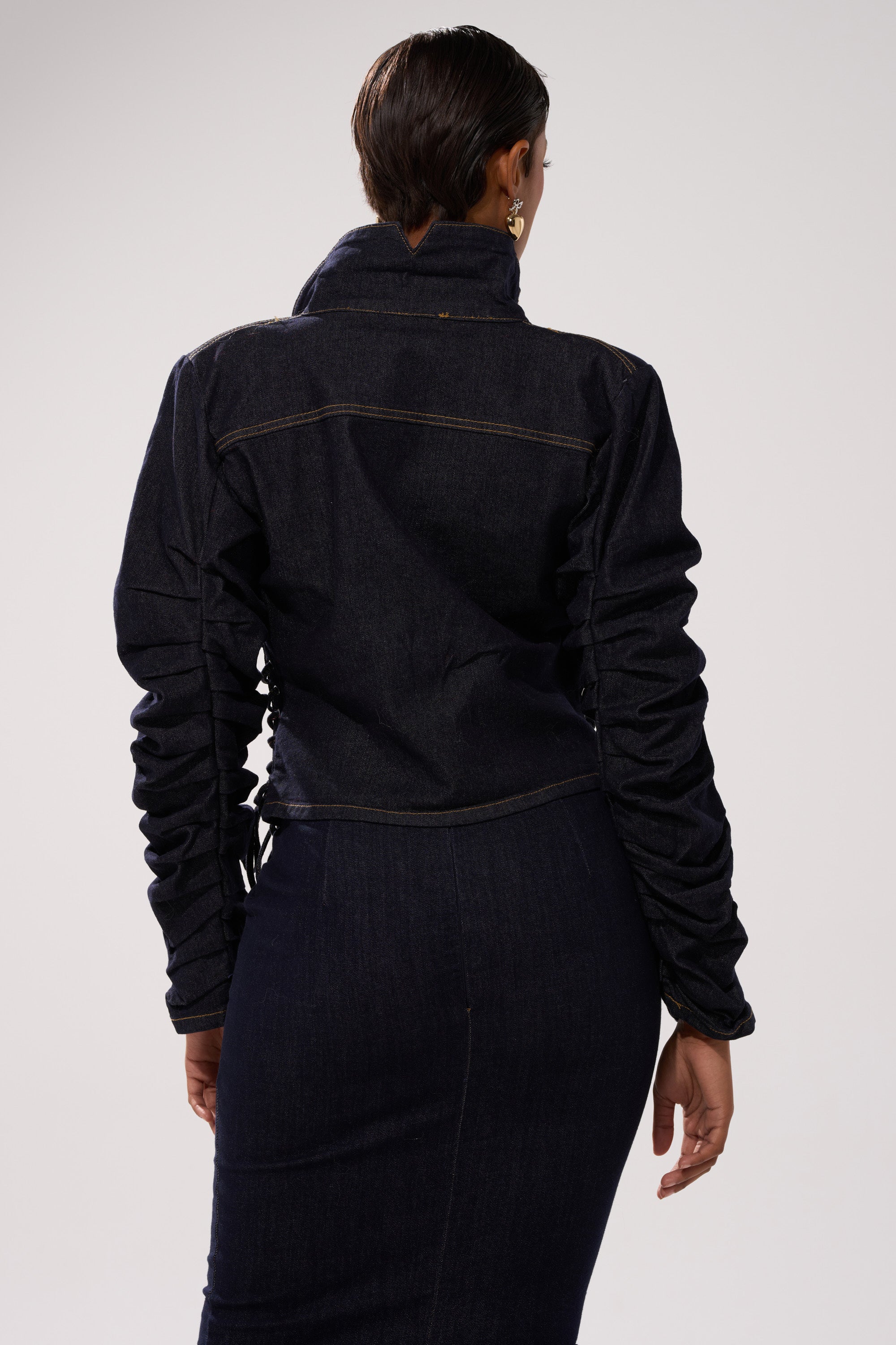 A person with short dark hair is seen from the back wearing the STRICTLY BUSINESS DENIM BLOUSE, a dark-wash denim top with ruched sleeves, paired with a matching high-waisted denim skirt against a plain light background.