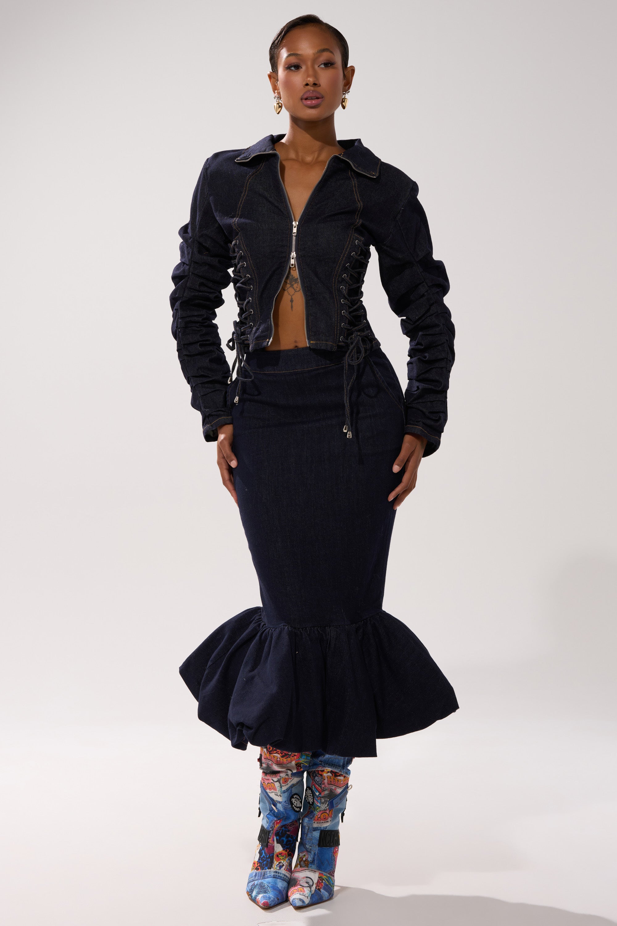 A woman models the STRICTLY BUSINESS DENIM BLOUSE, a dark-wash jacket with ruched sleeves, paired with a matching flared mermaid skirt, colorful patterned knee-high boots, and hoop earrings against a plain light background.