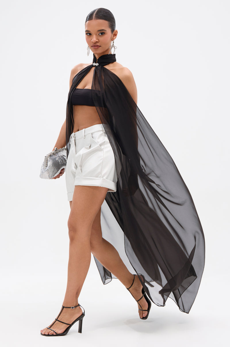 A woman walks confidently against a plain white background wearing DJ METALLIC SHORTS, a black bandeau top, a sheer black cape, heeled sandals, and holding a silver purse. 4