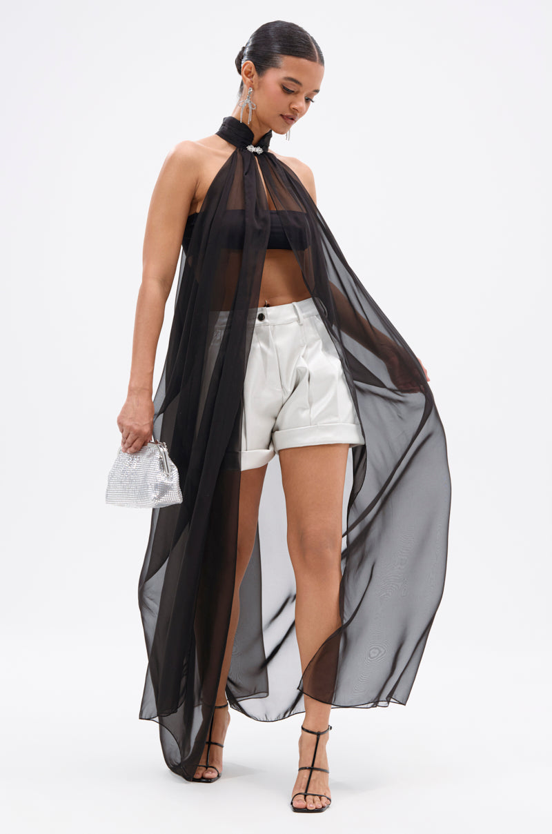 A woman wears a sheer black halter cape over a black bandeau top and DJ METALLIC SHORTS, holding a small silver purse with strappy black heels and her hair tied back. 5