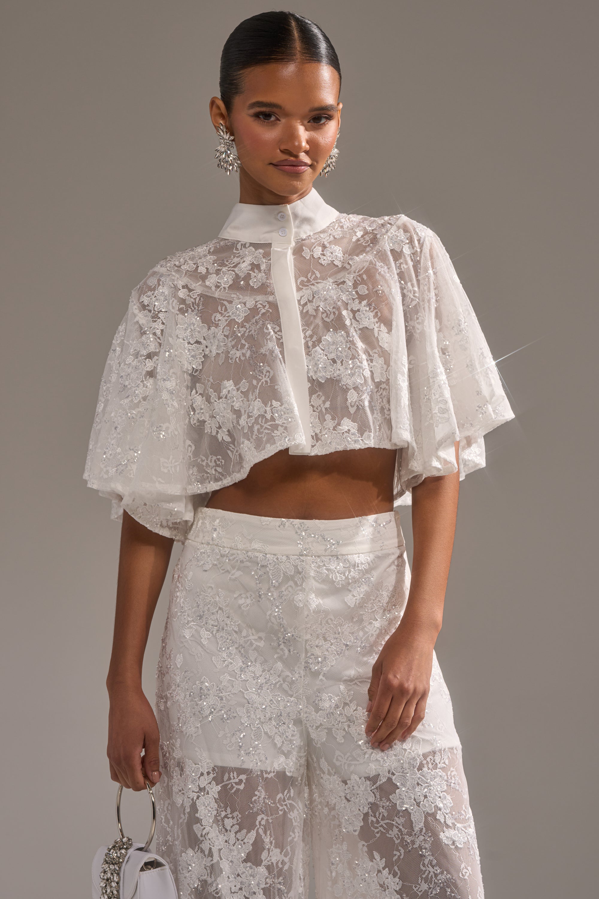 A woman wears the EVA LACE SHORT SLEEVE BUTTON DOWN with matching high-waisted lace pants, statement earrings, and a small white handbag, standing against a neutral background. 1