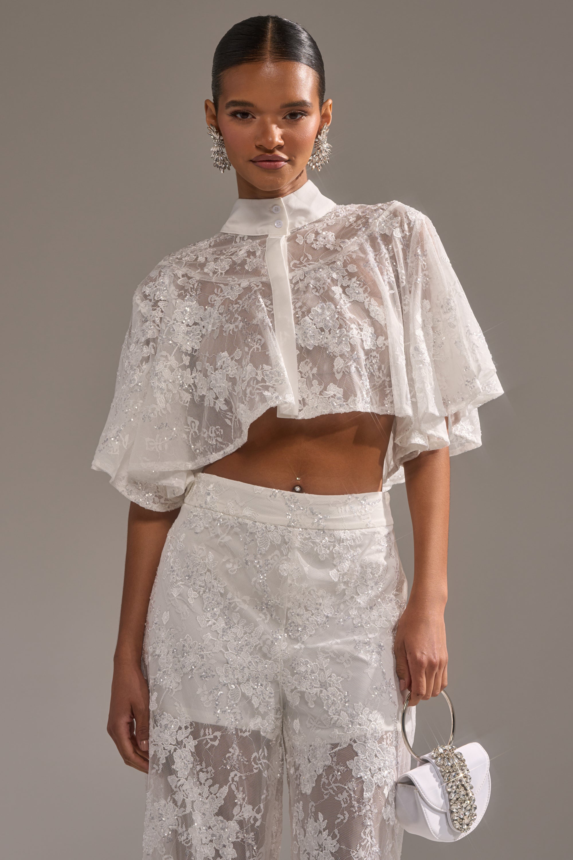 Someone wearing the EVA LACE SHORT SLEEVE BUTTON DOWN, featuring sheer white floral mesh with wide sleeves, paired with matching high-waisted lace pants, a small white chain-handle handbag, and large sparkling earrings. 2
