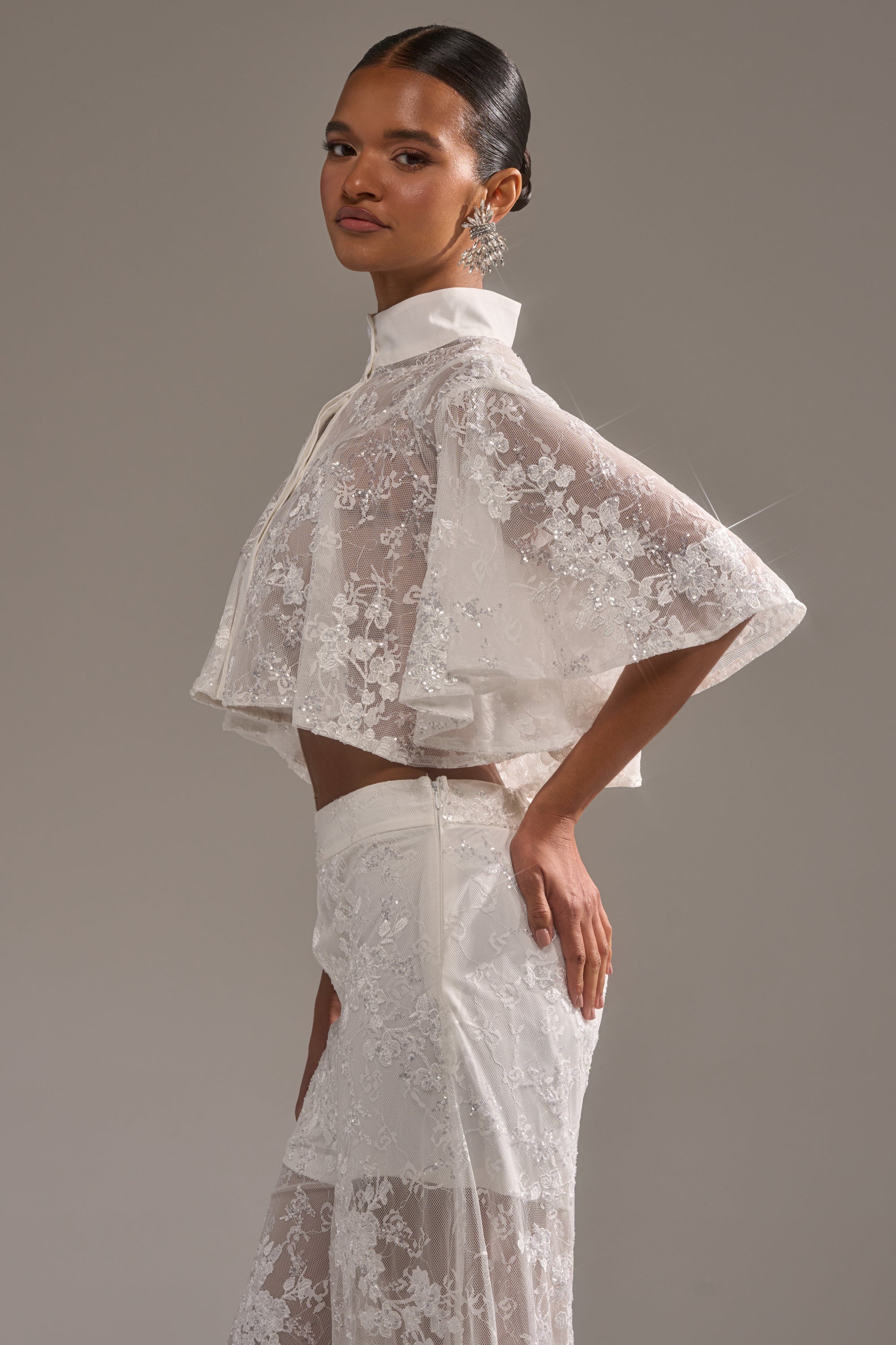 A woman models the EVA LACE SHORT SLEEVE BUTTON DOWN, a sheer white lace top with floral embroidery and sequins, paired with matching pants, set against a neutral background. 3