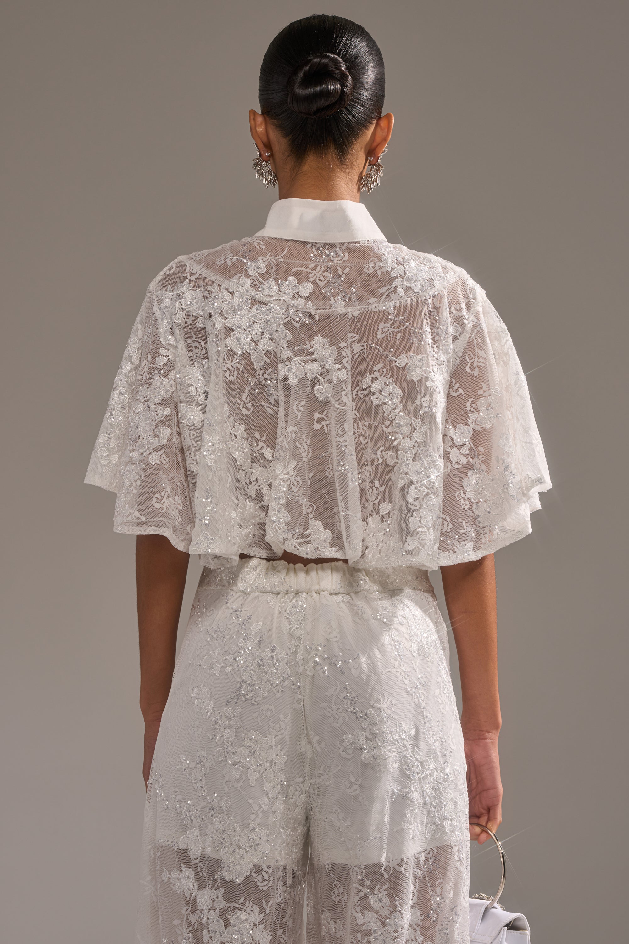 A person with dark hair in a sleek bun is seen from the back wearing the EVA LACE SHORT SLEEVE BUTTON DOWN, paired with wide-leg pants. They hold a small bag and accessorize with statement earrings. 4