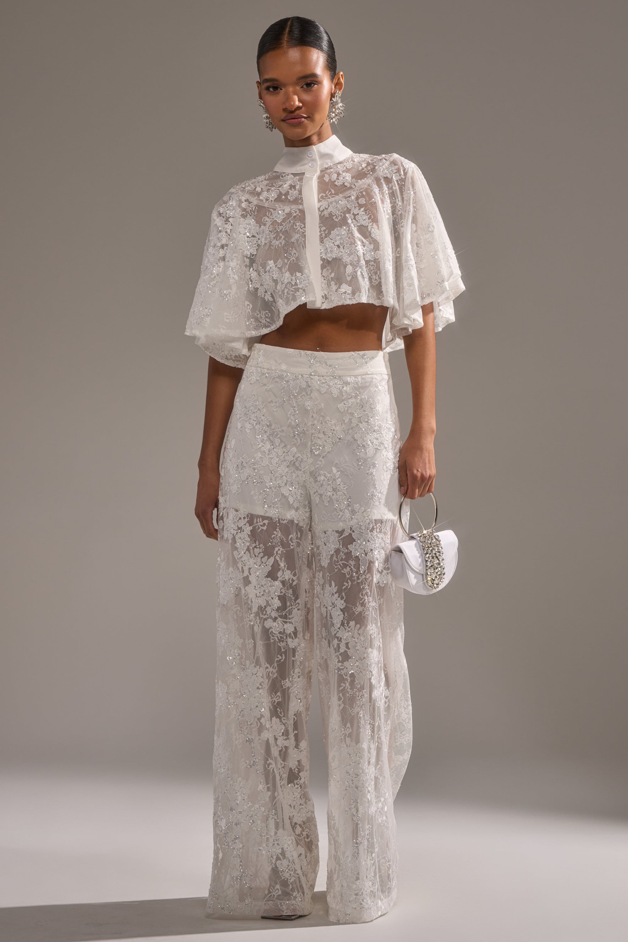 A woman wears the EVA LACE SHORT SLEEVE BUTTON DOWN, styled with high-waisted wide-leg pants and a small white handbag. Her hair is pulled back as she stands against a plain gray background. 5