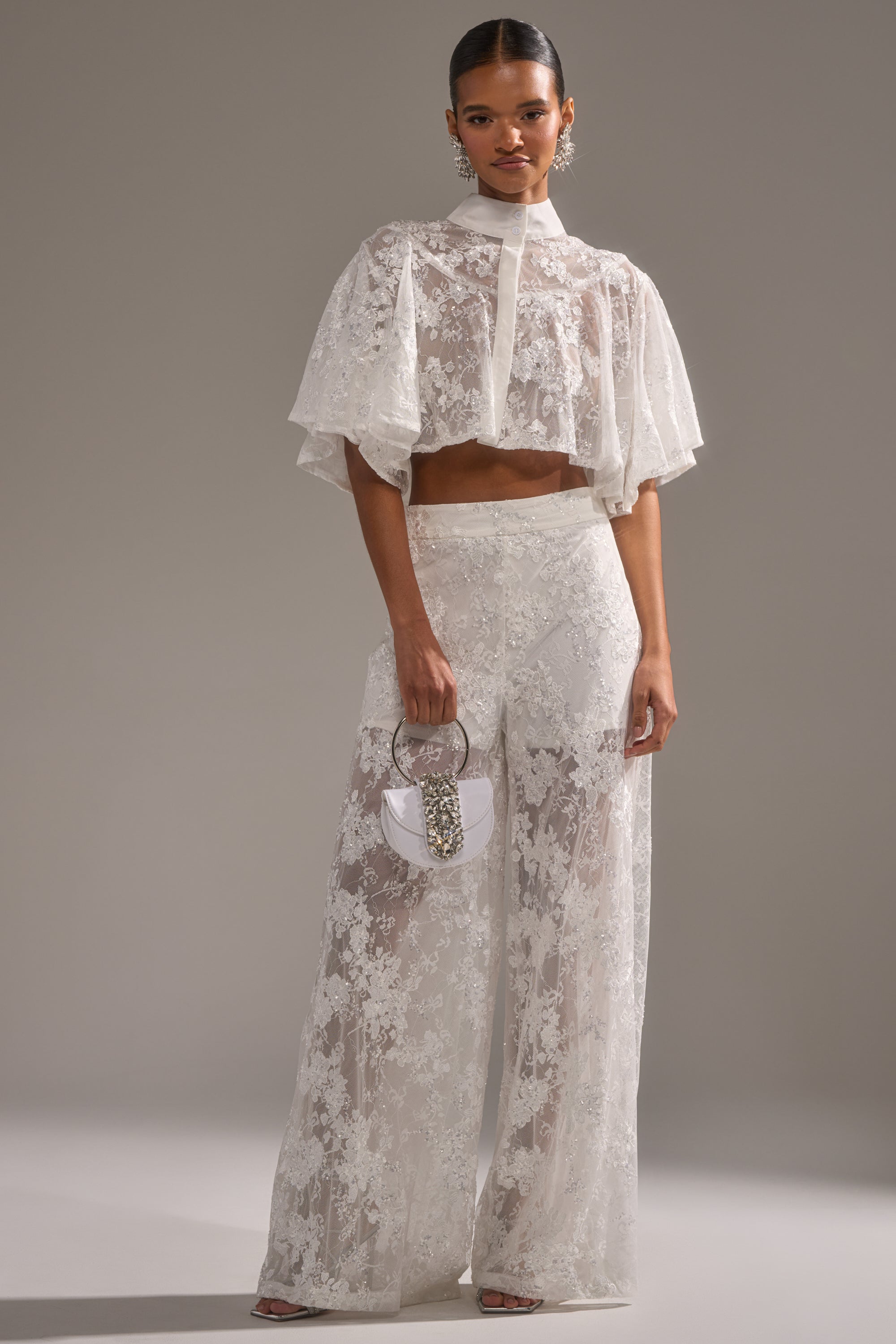 A woman stands in a studio wearing the EVA LACE SHORT SLEEVE BUTTON DOWN styled with sheer floral mesh wide-leg pants, a small white handbag, earrings, and slicked-back hair for a chic, modern look. 6