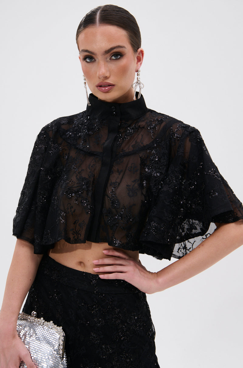 A woman wearing the QUEEN B LACE BUTTON DOWN with matching high-waisted pants holds a silver sequined clutch. She has slicked-back hair, drop earrings, and bold makeup against a plain light background.