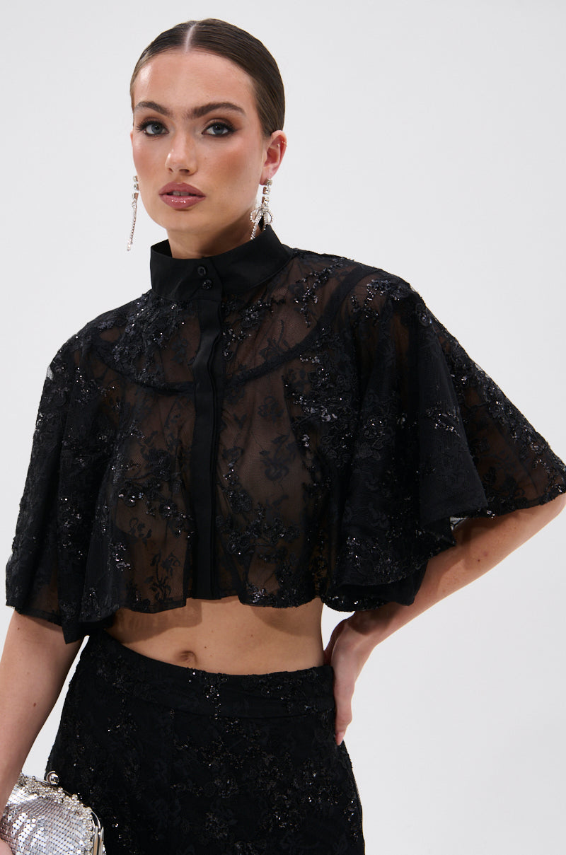 A woman wears the QUEEN B LACE BUTTON DOWN, a sheer black floral mesh cropped top with short sleeves and sequin accents, paired with matching high-waisted bottoms. She accessorizes with statement earrings and a sparkly silver clutch.